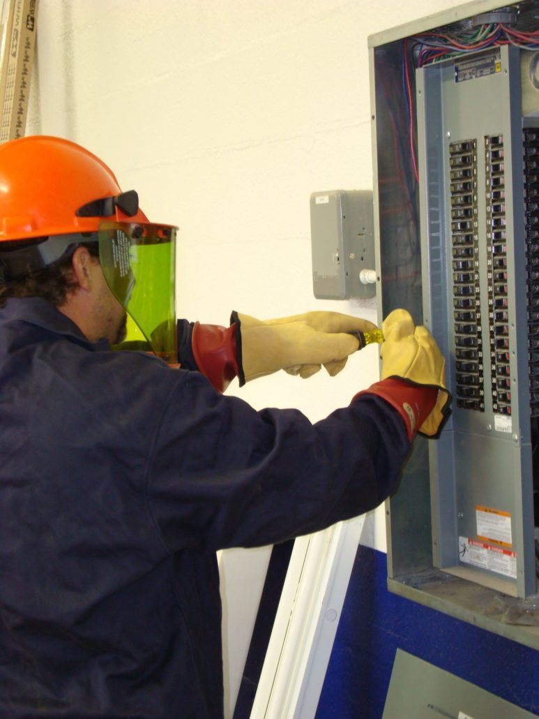 Electrical Orange hardhat Electrician wearing safety gloves & shield doing preventative maintenance work on a panel with a screwdriver