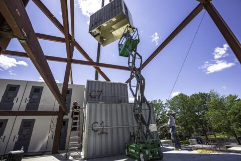 photo of a jail cell hoisted in the air as an electrical technician on a crane works below it.